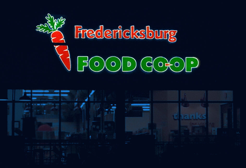 Illuminated channel letter sign for the Fredericksburg Food Co-Op at nighttime, featuring a prominent carrot icon, with the storefront visible below.