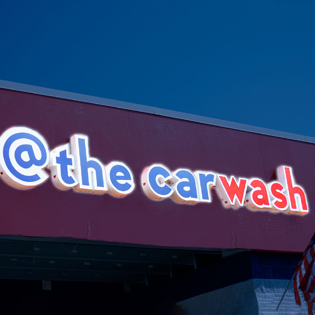 A brightly lit channel letter sign with the words '@ the carwash' in vibrant letters, illuminated on the front of a building.