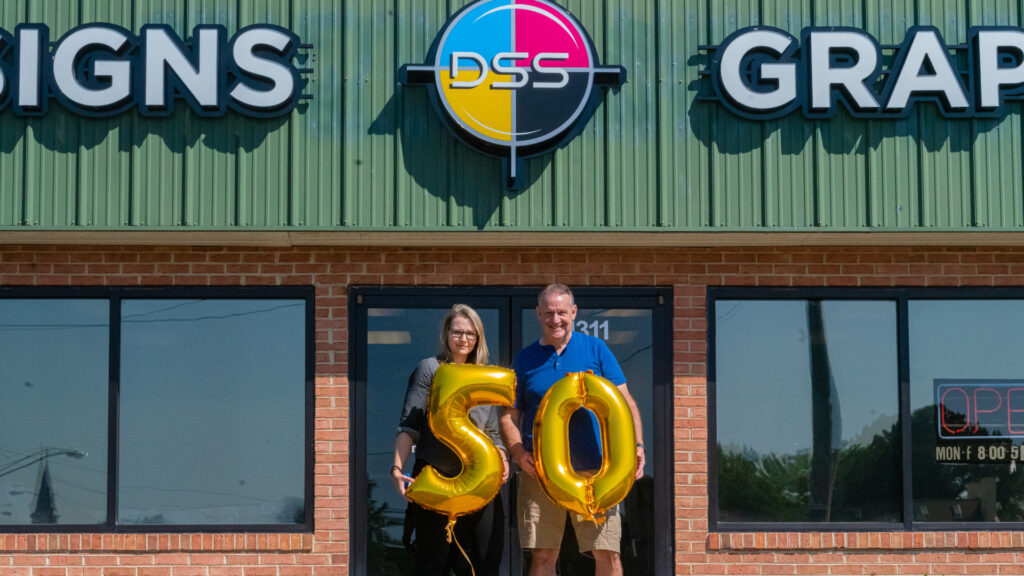 Distinct Signs President and Vice President stand in front of their building holding "50" balloons to celebrate the store's 50th anniversary. 