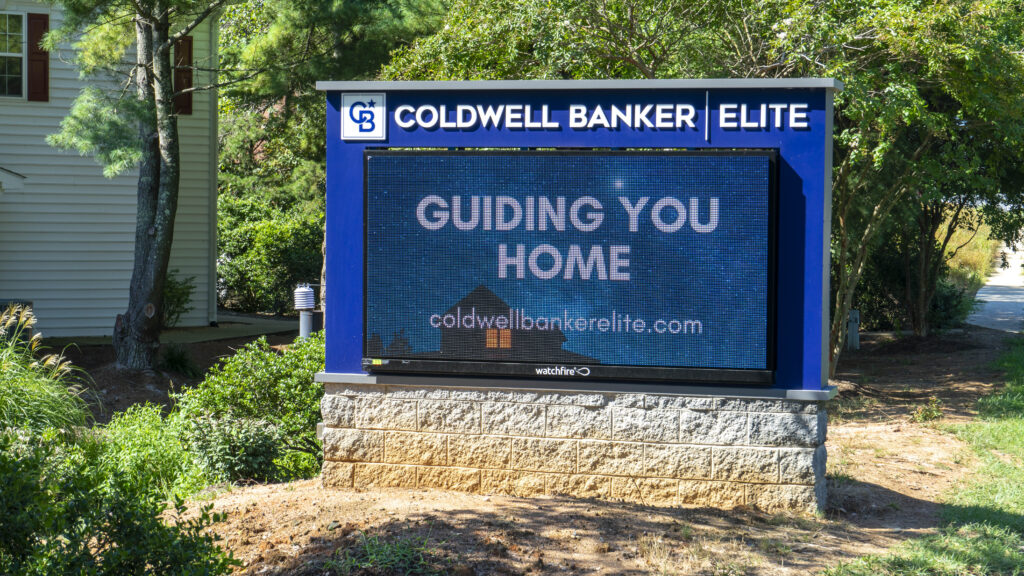 An electronic message board by Distinct Sign Solutions featuring Coldwell Banker Elite's branding and the message 'GUIDING YOU HOME,' set against a backdrop of greenery in Stafford, VA.