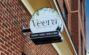 alt="Photo of Floral LED Illuminated blade sign displaying Veera Aesthetics mounted with black steel bracket on brick wall in Fredericksburg, Virginia"