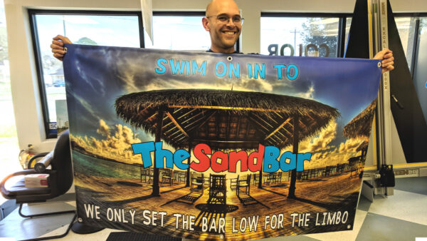 Cheerful man holding banner for The Sand Bar with tropical tiki hut and sunset background, created by Distinct Sign Solutions in Fredericksburg, VA.