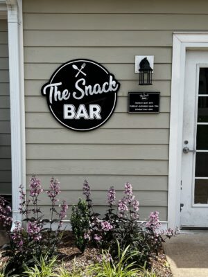 alt="Photo of Exterior Mounted Signs on tan siding for a restaurant called The Snack Bar. Sign is black and white and located in Fredericksburg, Virginia"