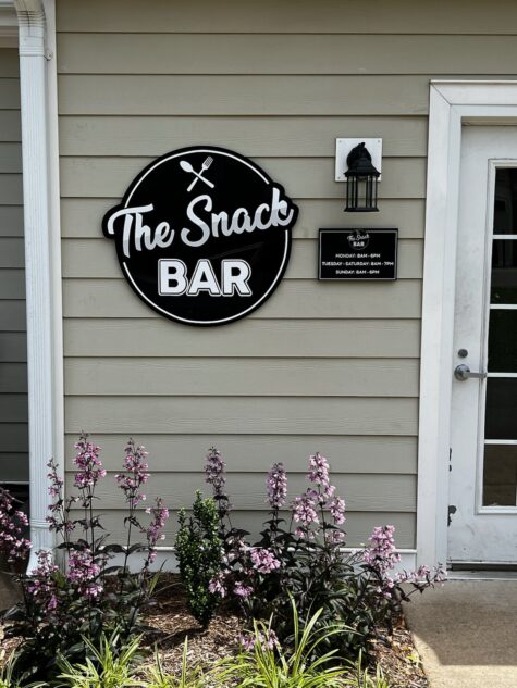 alt="Photo of Exterior Mounted Signs on tan siding for a restaurant called The Snack Bar. Sign is black and white and located in Fredericksburg, Virginia"