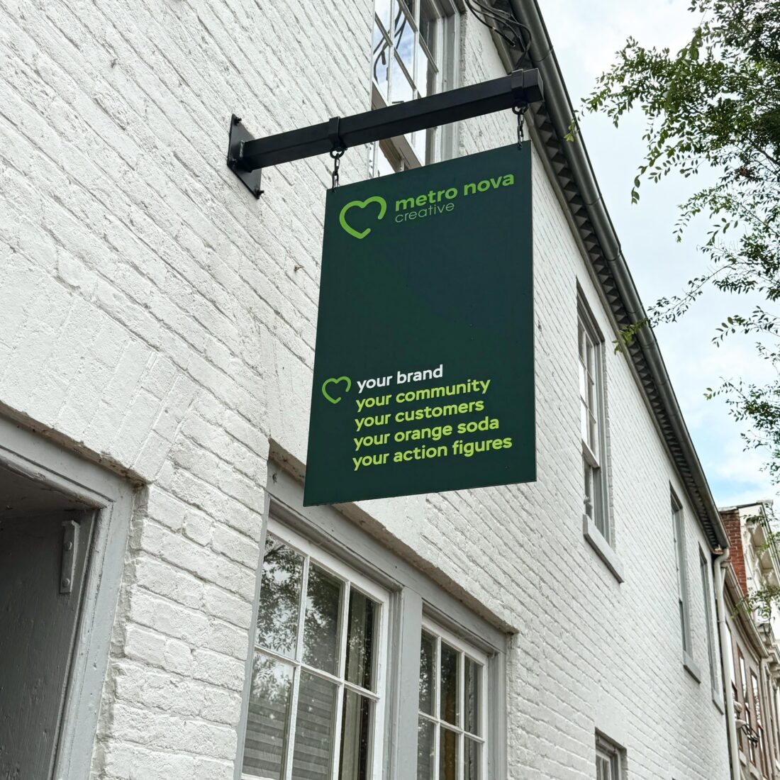 alt="Ground Photograph of blade sign hanging off of black steel mounted bracket. Sign is various shades of green and says metro nova creative: your brand, your community, your customers, your orange soda, your action figures in Fredericksburg, Virginia"