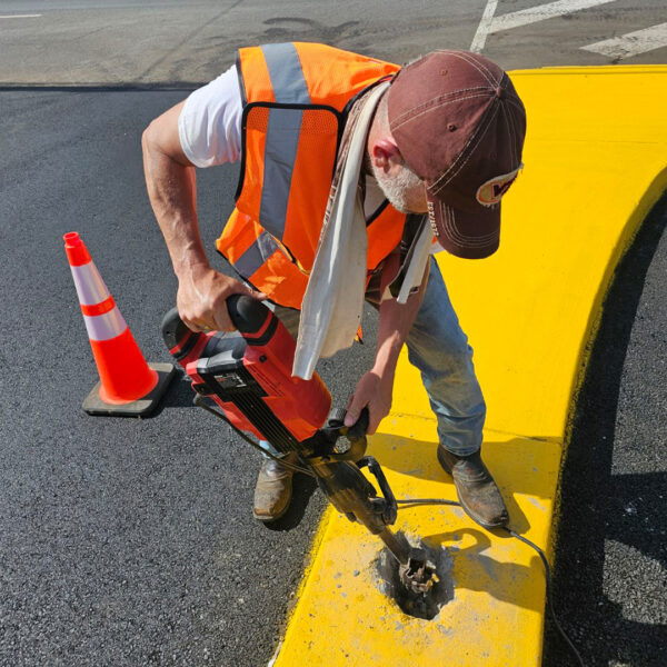 A local sign maker wearing a high-visibility orange vest and a brown cap uses a power drill to create a foundation hole for a directional sign in a bright yellow curb.