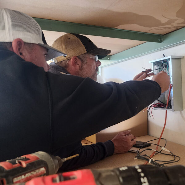 Two sign professionals in baseball caps carefully connect wiring inside a small electrical panel, preparing the circuitry that will illuminate a sign.