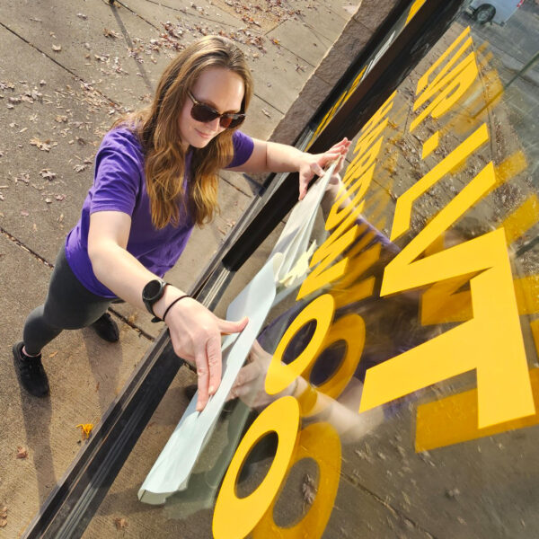 A vinyl specialist carefully applies bright yellow lettering to a storefront window, ensuring each character is perfectly placed and secure.