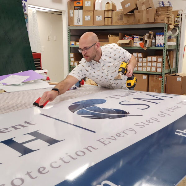 A local sign maker wearing glasses and a patterned shirt carefully smooths a large acrylic tenant panel in a workshop, surrounded by tools and boxes.