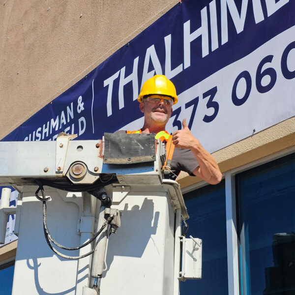 A worker in a yellow hard hat and orange safety vest stands in a bucket truck lift, giving a thumbs-up as he positions a large vinyl banner on a building’s exterior.