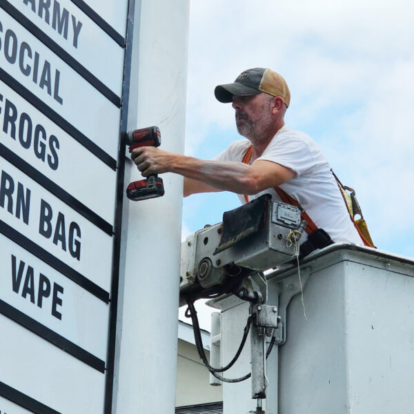 A local sign maker in a baseball cap uses a cordless drill to fasten a panel onto a freestanding pylon sign while working from a raised bucket truck.