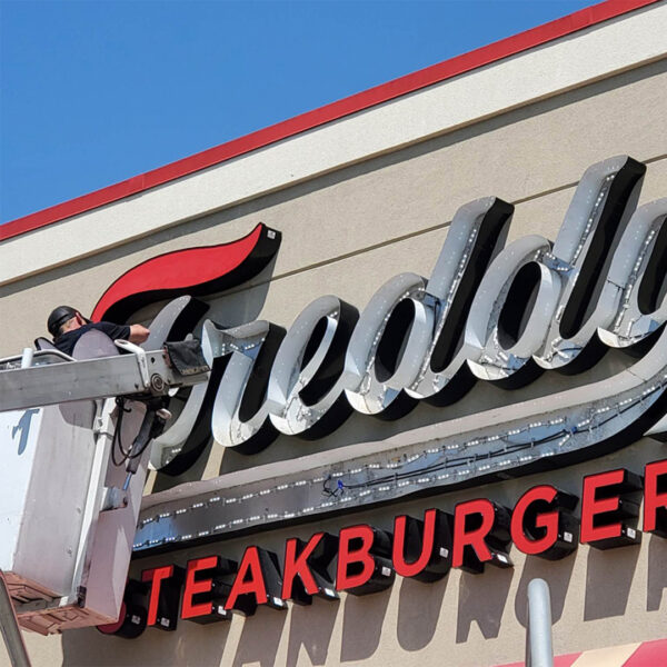 A Distinct Sign Solutions team member in a bucket truck aligns and secures the large, three-dimensional letters of a building-mounted “Freddy’s Steakburger” sign during a sign repair.