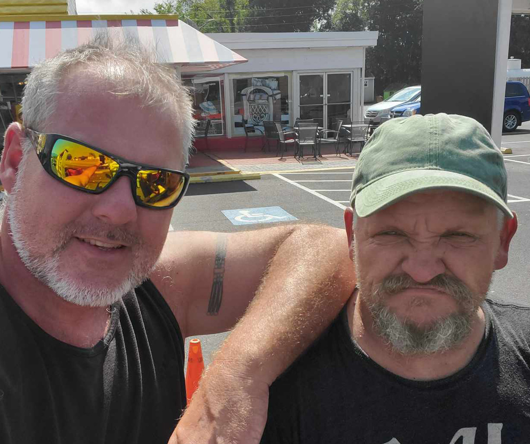 Two local sign makers—one grinning in reflective sunglasses and the other making a playful, scrunched-up face—pose together in a parking lot with a building in the background.