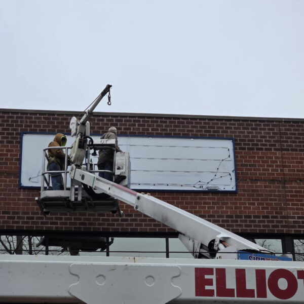 Installers in bucket truck mounting UL-Listed cabinet sign frame for Atlantic Builders on brick storefront.