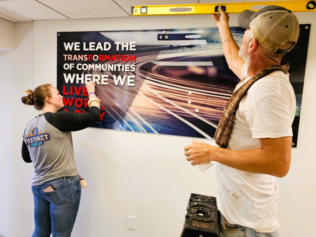 Photo of two installers doing finishing touches on interior acrylic wall sign in Fredericksburg, VA. Sign was manufactured & installed by Distinct Sign Solutions in Fredericksburg, VA
