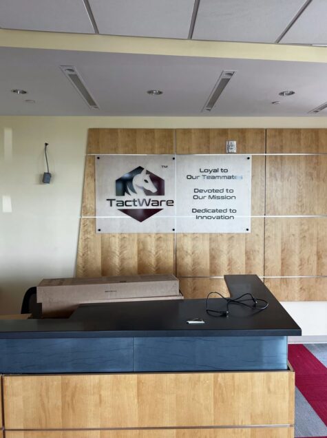 Photo of frosted acrylic with black & burgundy TactWare logo set to wooden receptionist wall in Stafford, VA. Sign was manufactured & installed by Distinct Sign Solutions in Fredericksburg, VA