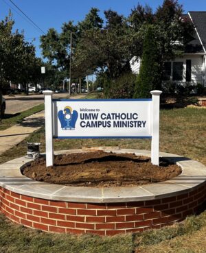 Photo of custom post & panel sign affixed to vinyl pvcwhite posts in brick base. Sign is for University of Mary Washington Catholic Campus Ministry , located in Fredericksburg, VA. Sign was manufactured & installed by Distinct Sign Solutions in Fredericksburg VA