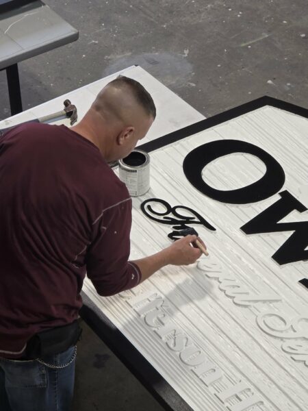 Sign craftsman hand-painting raised dimensional lettering on a carved commercial sign panel during fabrication.
